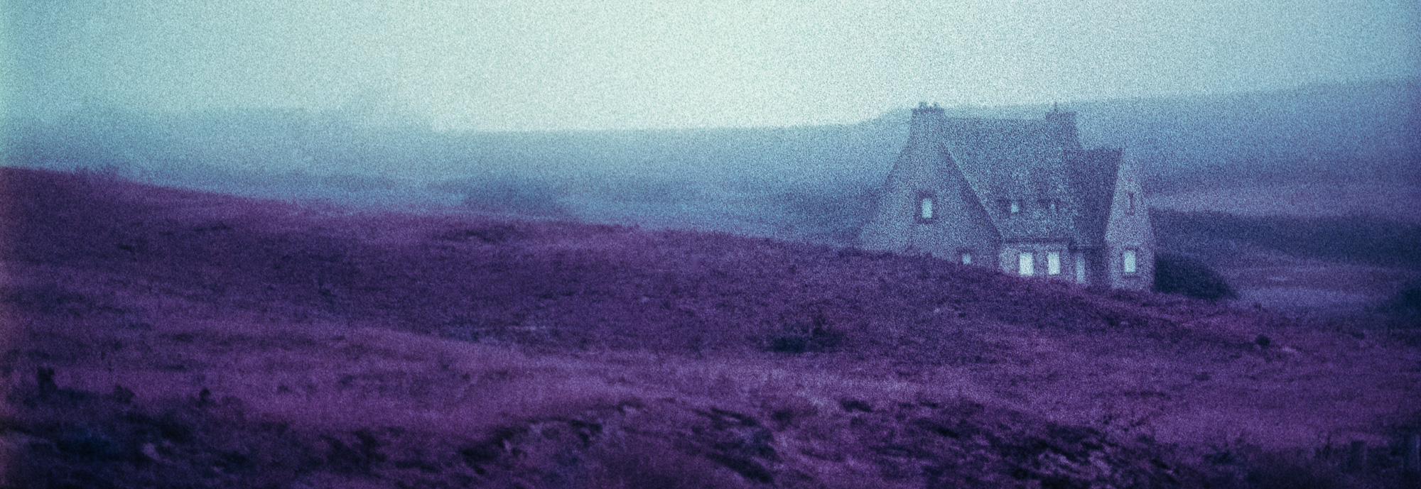 Photography of hills and a stone building in Brittany. The colors of the photography are reversed: the hills are strong purple, the sky appears turquoise.